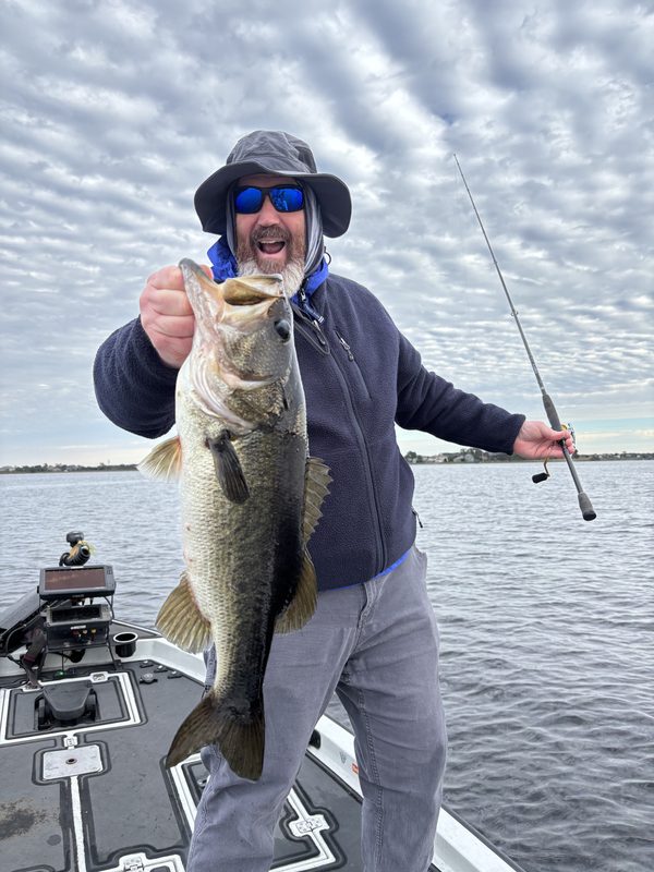 Angler in a sun hat holding up a largemouth bass and a spinning rod on a cloudy winter day