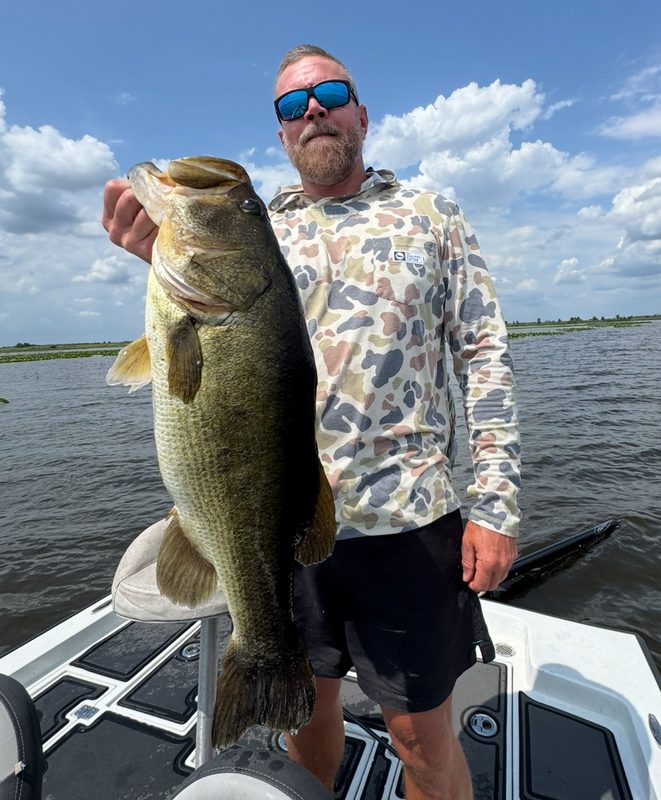 Angler in a camo performance shirt holding a large largemouth bass on a bass boat deck