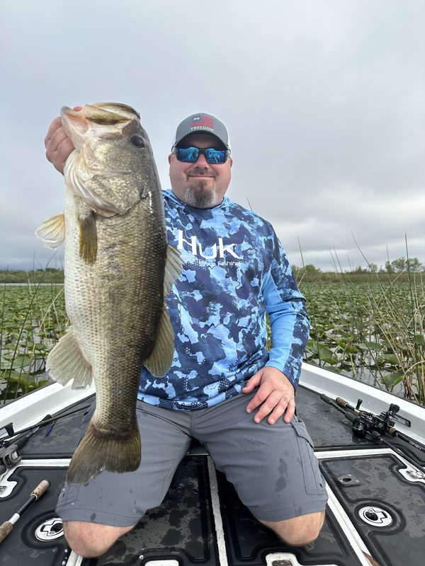 Angler kneeling on a bass boat deck holding a big largemouth bass beside a lily pad field