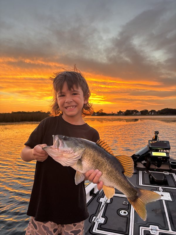 Young angler holding a largemouth bass against an orange sunset from a bass boat
