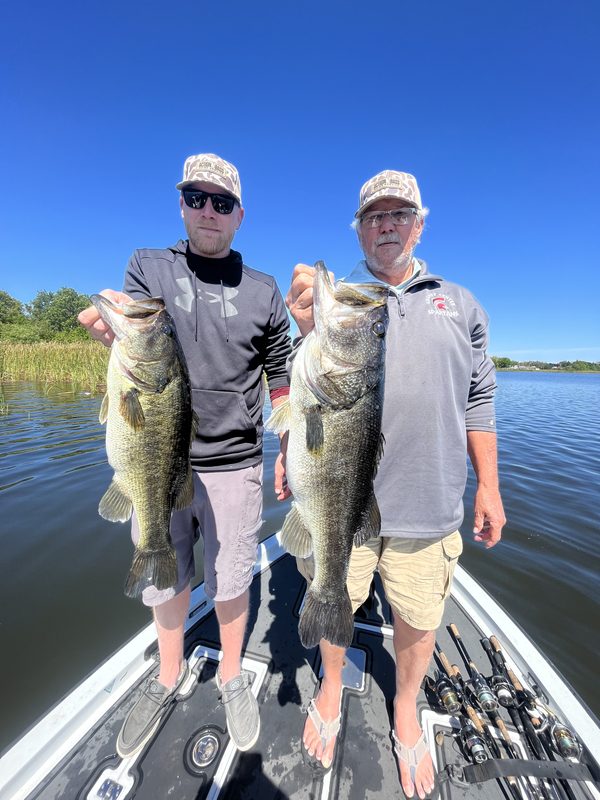 Two anglers standing on a bass boat bow each holding a largemouth bass beside a reed line, Winter Haven Chain of Lakes